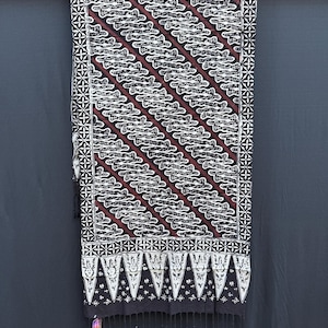 Peut inclure: Un textile rectangulaire présentant un motif batik diagonal répétitif en noir, blanc et bordeaux foncé. Le tissu est bordé d'un motif géométrique en étoile et fini avec un bord inférieur composé de formes triangulaires, de fleurs et de franges.