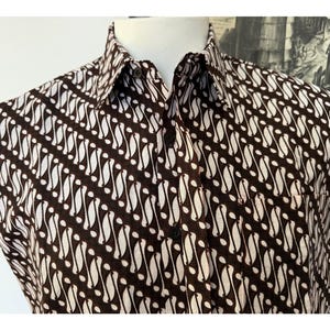 Pode incluir: Uma camisa de manga curta com botões e um padrão batik diagonal em castanho escuro e branco sujo. A peça apresenta um colarinho clássico pontiagudo, uma carcela frontal com botões escuros e um bolso no peito no lado esquerdo.