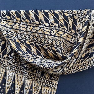 Peut inclure: Un tissu noir et beige présentant un motif batik détaillé avec des formes géométriques répétitives, des motifs floraux et des lignes ondulées. L'étoffe est drapée sur une barre horizontale, affichant un agencement complexe de bordures.
