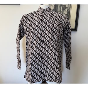 May include: A long-sleeved button-down shirt featuring a dark brown and white diagonal batik pattern. The shirt has a standard collar, a front button placket, and long sleeves with cuffs, displayed on a mannequin against a plain white wall.