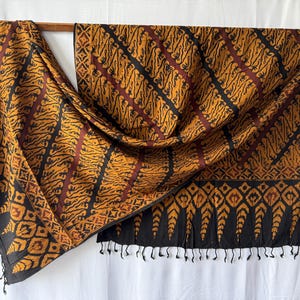 Pode incluir: Um têxtil retangular com um padrão batik repetitivo em tons de amarelo mostarda, castanho escuro e preto. O tecido apresenta riscas diagonais com motivos orgânicos intrincados, uma borda geométrica e uma extremidade com franjas pretas.