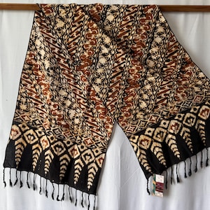 May include: A rectangular textile wrap featuring a brown, black, and cream batik pattern with diagonal stripes and floral motifs. The fabric has a decorative border with geometric shapes and a fringe of black tassels along the bottom edge.