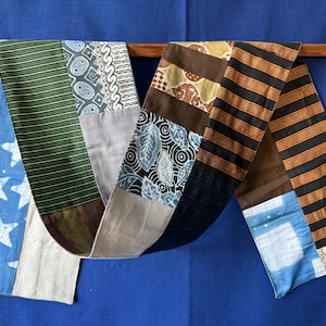 Peut inclure: Un long chemin de table rectangulaire composé de différents motifs textiles en patchwork. Les sections présentent du tissu bleu à étoiles blanches, des rayures vertes et jaunes, des imprimés géométriques gris, des motifs de feuilles noir et blanc, des panneaux marron et des rayures orange et noires.