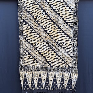 Peut inclure: Un textile rectangulaire présentant un motif batik noir et crème avec des bandes diagonales de formes organiques stylisées. Le tissu possède une bordure décorative avec des motifs en croix et un bord inférieur avec des triangles et des franges.