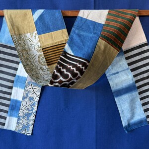 Peut inclure: Une longue écharpe rectangulaire présentant un motif patchwork composé de divers tissus. Les motifs incluent des rayures horizontales noires et blanches, du tie-dye bleu et blanc, des imprimés batik floraux, des sections beige uni et du tissu rayé vert foncé et marron.
