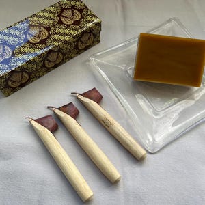 Pode incluir: Três canetas de caligrafia de madeira com pontas marrons e vermelhas, uma pequena caixa com um design de padrão marrom e dourado e um prato de vidro transparente com um bloco de cera marrom.