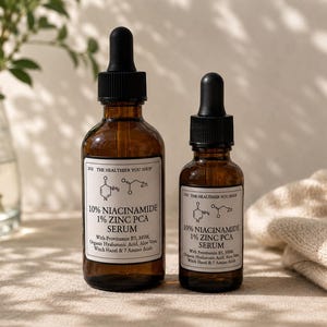 May include: Two amber glass serum bottles with black droppers. The bottles have white labels with text including "10% Niacinamide 1% Zinc PCA Serum". The background includes a plant in a glass vase and a folded beige cloth.
