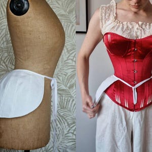 May include: A white, curved underbust corset with tie fastenings is displayed on a mannequin. A red satin corset with vertical white accents is worn with white trousers. The garments are examples of historical fashion.