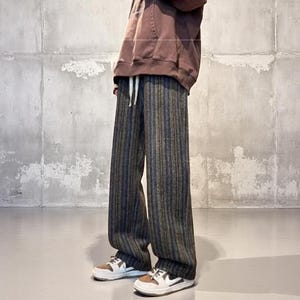 May include: Wide-leg pants with vertical stripes in shades of gray, blue, and brown. The pants have a drawstring waist and are paired with white and brown sneakers. The person is wearing a brown hoodie.