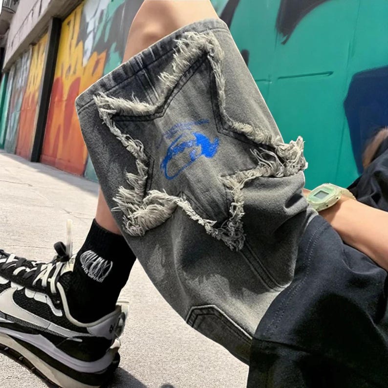May include: Gray denim shorts with a distressed star design and blue graphic. The shorts have frayed edges and a faded appearance. The person is wearing black socks with white text and black and white sneakers.