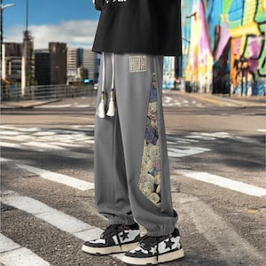 May include: Gray sweatpants with a colorful graphic print on the side. The sweatpants have a drawstring waistband and a small embroidered logo on the front.