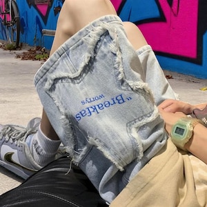 May include: Light blue denim shorts with distressed edges and the embroidered text "Breakfast Worries" in blue. The shorts are worn with a light-colored top and sneakers. A watch is visible on the wrist.