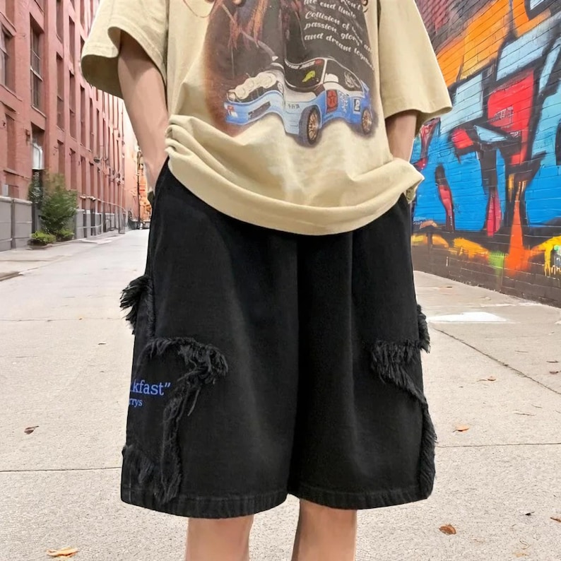 May include: Black denim shorts with distressed detailing and pockets. The shorts have a relaxed fit and feature the text "kfast" in blue on one pocket. The shorts are worn with a tan t-shirt.