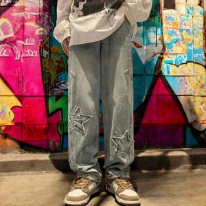 Könnte beinhalten: Hellblaue Jeans aus Denim mit großen gestickten Sternen. Die Jeans sind locker geschnitten und haben ein gerades Bein.