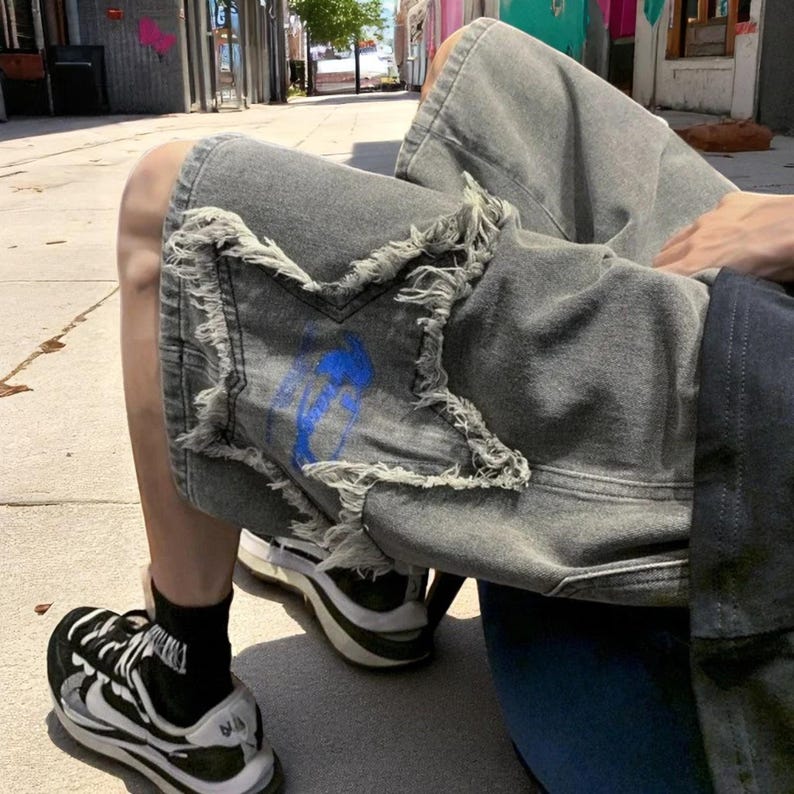 May include: Gray denim shorts with distressed edges and a blue painted design. The shorts are worn with black and white sneakers and black socks. The overall style is casual and urban.