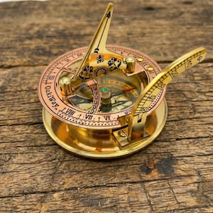 May include: A vintage brass sundial compass with a copper-colored ring and gold accents. The compass features Roman numerals, a needle, and a gnomon. The sundial is set on a wooden surface.