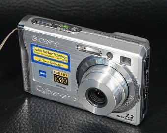 May include: Silver Sony Cyber-shot DSC-W80 digital camera with a Carl Zeiss lens. Features include Full HD 1080, 7.2 megapixels, and Double Anti-blur Technology. The camera has a retractable lens and a small flash.