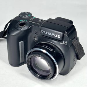 May include: Black Olympus SP-500UZ digital camera with a zoom lens. The camera has a 6.0 megapixel resolution and a black strap. The lens has the text "AF ZOOM 6.3-63mm 1:2.8-3.7".