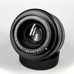 May include: A black Leitz Canada Summicron-R 1:2/35 camera lens with a serial number 2615145 printed on the front ring. The lens features a textured focus ring, a glass front element, and is positioned on top of a black circular lens cap.