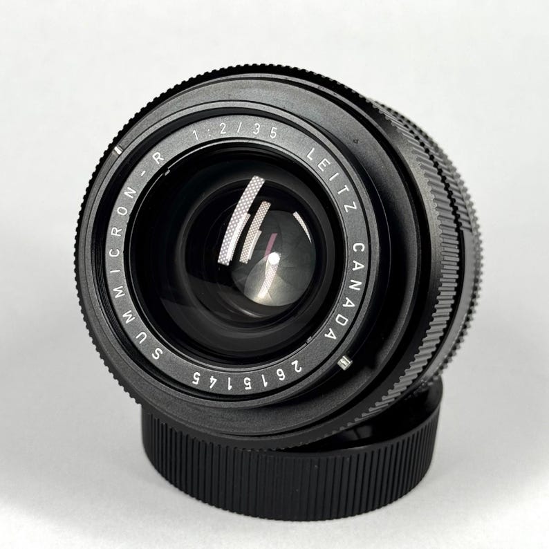 May include: A black Leitz Canada Summicron-R 1:2/35 camera lens with a serial number 2615145 printed on the front ring. The lens features a textured focus ring, a glass front element, and is positioned on top of a black circular lens cap.
