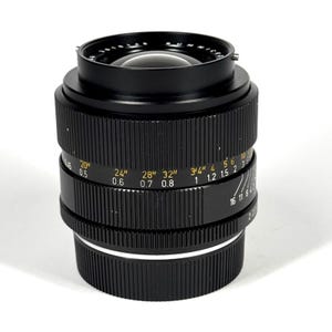 May include: A black camera lens featuring a ridged focus ring with yellow distance markings in inches and white markings in meters. The lens barrel displays aperture settings and the text SUMMICRON, with a glass element visible at the top.