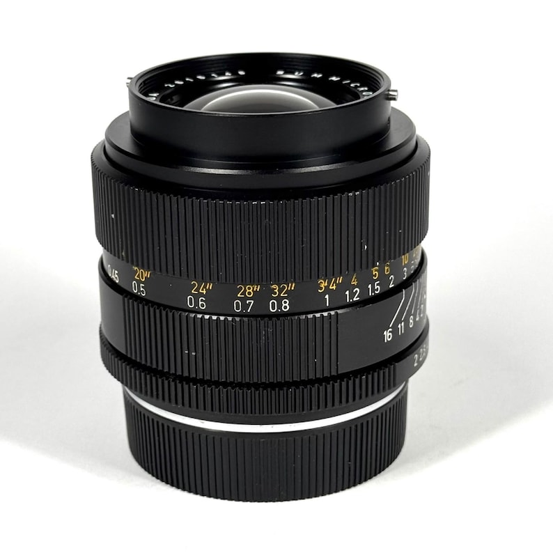May include: A black camera lens featuring a ridged focus ring with yellow distance markings in inches and white markings in meters. The lens barrel displays aperture settings and the text SUMMICRON, with a glass element visible at the top.