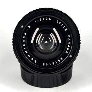May include: A black Leitz Canada Summicron-R 1:2/35 camera lens with a textured focus ring and a smooth front element. The lens barrel features white engraved text reading Summicron-R, 1:2/35, Leitz Canada, and the serial number 2615145.