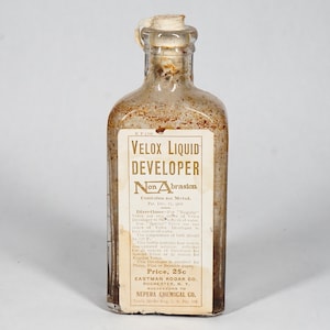May include: A vintage glass bottle of Velox Liquid Developer, a photographic chemical. The label is yellow with black text and reads "Velox Liquid Developer Non Abrasion" and "Contains no Metol." The bottle is partially filled with a clear liquid.