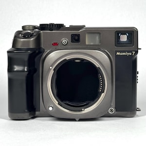 May include: A Mamiya 7 medium format rangefinder camera body in a metallic gray finish. The front features a large circular lens mount opening, a black grip on the left side, a viewfinder window, and the Mamiya 7 logo printed in gold on the right.