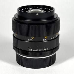 May include: A black camera lens with a ribbed focus ring and a textured aperture ring. The barrel features yellow markings for distance in inches and white markings for distance in meters, with the text LENS MADE IN CANADA printed on the side.