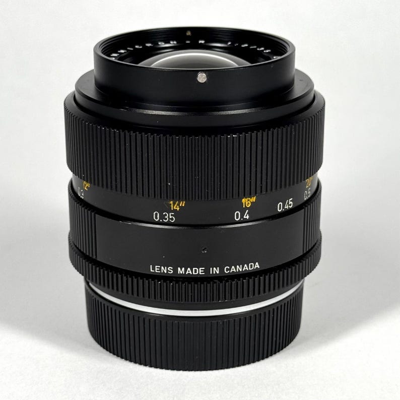May include: A black camera lens with a ribbed focus ring and a textured aperture ring. The barrel features yellow markings for distance in inches and white markings for distance in meters, with the text LENS MADE IN CANADA printed on the side.