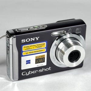 May include: A black Sony Cyber-shot digital camera with a silver Carl Zeiss Vario-Tessar lens. The front features a sticker with text including Double Anti-blur Technology, Face Detection, and Full HD 1080. The top has a silver shutter button.