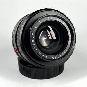 May include: A black Leitz Canada Summicron-R 1:2/35 camera lens with a serial number 2615145 printed on the front ring. The lens features a textured focus ring, a red alignment dot on the side, and a black circular lens cap at the base.