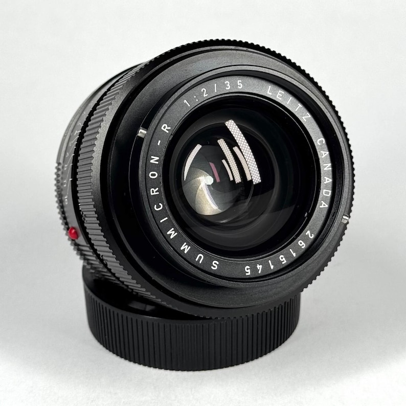 May include: A black Leitz Canada Summicron-R 1:2/35 camera lens with a serial number 2615145 printed on the front ring. The lens features a textured focus ring, a red alignment dot on the side, and a black circular lens cap at the base.