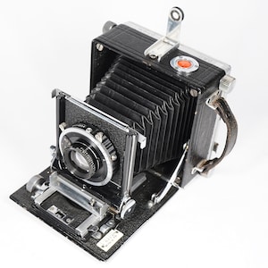 May include: A black and silver vintage camera with a bellows system and a lens with a black and silver lens cap. The camera has a leather strap attached to it.