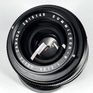 May include: A black Leitz Canada Summicron-R 1:2/35 camera lens with a circular front element. The lens barrel features white engraved text reading 2615145, SUMMICRON-R, 1:2/35, and LEITZ CANADA. The internal aperture blades are visible inside.