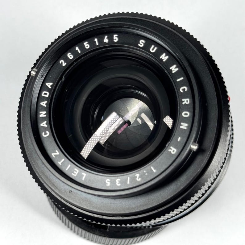 May include: A black Leitz Canada Summicron-R 1:2/35 camera lens with a circular front element. The lens barrel features white engraved text reading 2615145, SUMMICRON-R, 1:2/35, and LEITZ CANADA. The internal aperture blades are visible inside.