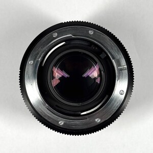 May include: A black camera lens mount viewed from the rear, featuring a circular metal flange with silver-colored screws, a black ridged focus ring, and a dark glass element reflecting a purple grid pattern in the center of the aperture.