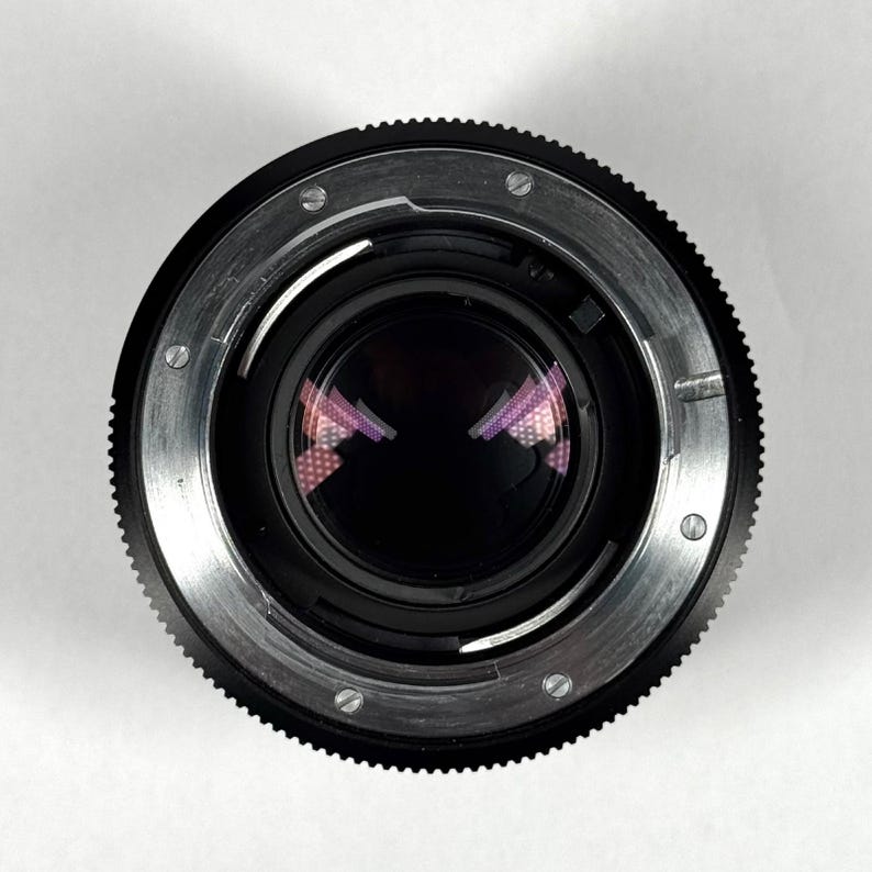 May include: A black camera lens mount viewed from the rear, featuring a circular metal flange with silver-colored screws, a black ridged focus ring, and a dark glass element reflecting a purple grid pattern in the center of the aperture.