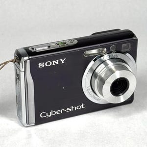 May include: A black Sony Cyber-shot digital camera with a silver Carl Zeiss Vario-Tessar lens. The camera features a rectangular body, a top-mounted shutter button, a built-in flash, and a thin brown wrist strap attached to the side of the device.