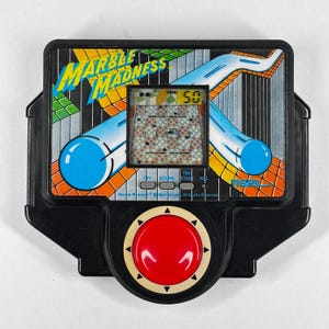 May include: A retro handheld electronic game, "Marble Madness," in a black plastic casing. The game has a small screen showing a maze, with a red button below. The game's logo is in yellow and blue.