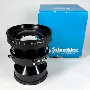 Op de afbeelding: Een zwarte Schneider Kreuznach Symmar-S 8.4/480 cameralens met een blauwe doos op de achtergrond. De lens heeft verschillende wijzerplaten en instellingen. De doos heeft het Schneider Kreuznach logo.