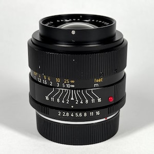 May include: A black camera lens with a ribbed focus ring and a manual aperture ring featuring white markings. The barrel displays distance scales in feet and meters in yellow, along with depth of field indicators and a small red alignment dot.