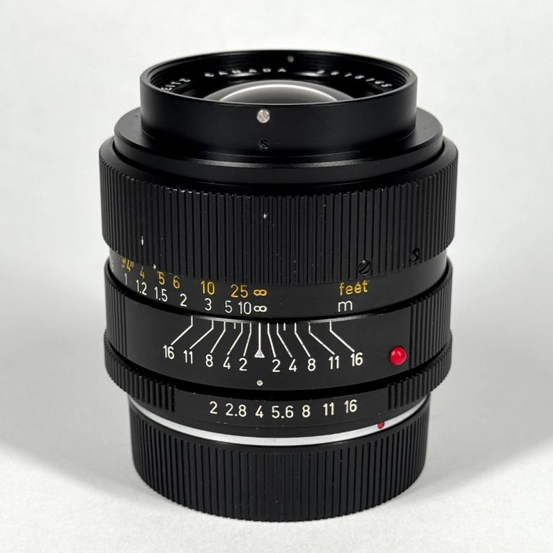 May include: A black camera lens with a ribbed focus ring and a manual aperture ring featuring white markings. The barrel displays distance scales in feet and meters in yellow, along with depth of field indicators and a small red alignment dot.