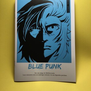 Peut inclure: Une brochure blanche avec une illustration bleue et noire d'un visage. Le titre "BLUE PUNK" est affiché en dessous. Du texte en français est également présent. La brochure est posée sur une surface jaune.