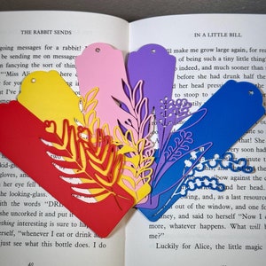 May include: Five colourful paper bookmarks with floral designs. The bookmarks are in shades of red, pink, yellow, blue, and purple. The bookmarks are placed on an open book with text.