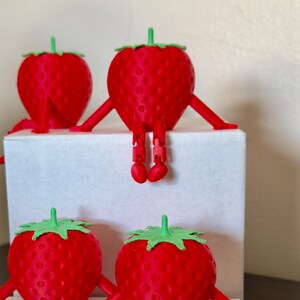 May include: Four red strawberry figurines with green tops and articulated arms and legs. Two strawberries sit on top of a white box, and two sit on the bottom. The strawberries are a whimsical, playful design.