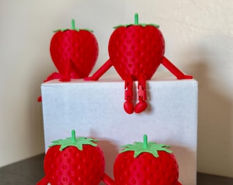 May include: Four red strawberry figurines with green tops and articulated arms and legs. Two strawberries sit on top of a white box, and two sit on the bottom. The strawberries are a whimsical, playful design.