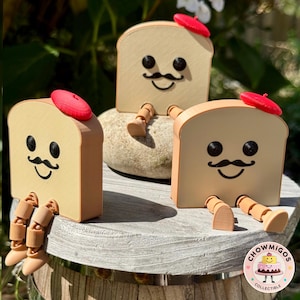 May include: Three whimsical bread slice figurines with smiling faces, mustaches, and red berets. The bread slices have articulated legs and are sitting on a stone and a wooden surface. The image is taken outdoors in natural light.