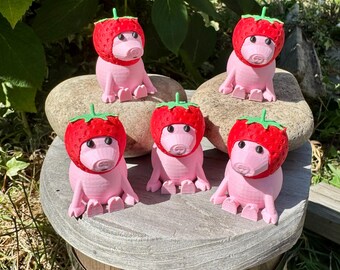 May include: Five pink pig figurines wearing red strawberry hats with green stems. The figurines are sitting on a wooden surface and a large rock. The background features green foliage and sunlight.